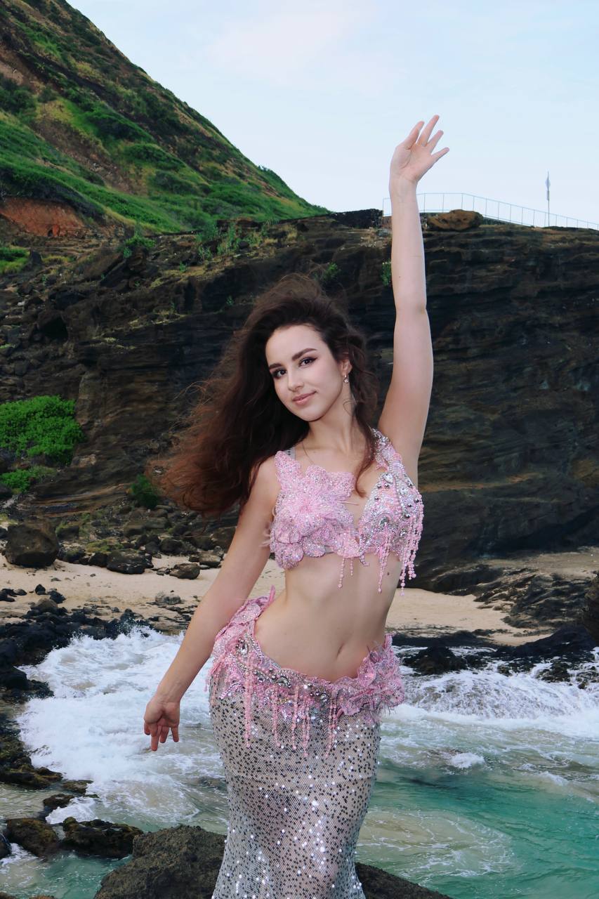 Angelina with one arm raised in a pink mermaid costume by the ocean